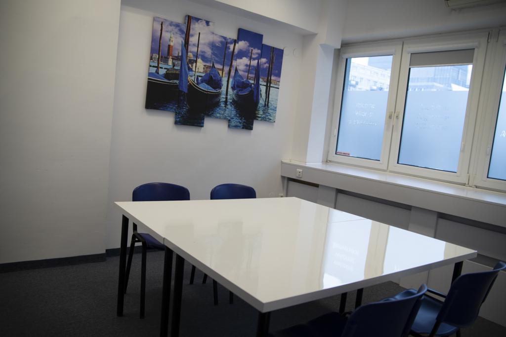Jasne biuro z białym stołem konferencyjnym otoczonym niebieskimi krzesłami, na ścianie wisi obraz przedstawiający weneckie gondole, okna z naklejkami informacyjnymi.
