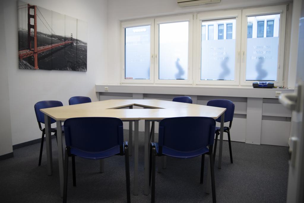 Sala konferencyjna z sześciokątnym stołem i niebieskimi krzesłami, na ścianie obraz mostu Golden Gate, okna z naklejonymi napisami w wielu językach.