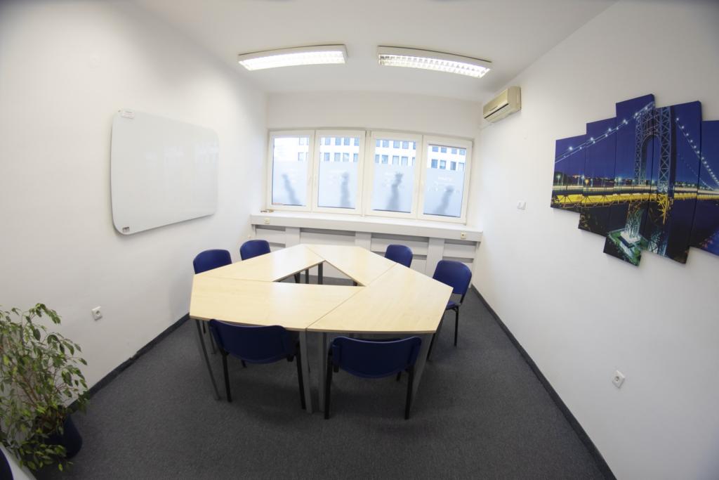 Jasna sala konferencyjna z trapezoidalnym stołem, niebieskimi krzesłami, białą tablicą i obrazem mostu nocą na ścianie.