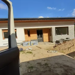 Nowoczesny dom jednorodzinny w trakcie prac wykończeniowych elewacji, widoczne częściowo ocieplone ściany, paczki styropianu Austrotherm przed wejściem, otwarte drzwi samochodu dostawczego w kadrze.