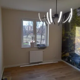 Pokój po remoncie: drewniana podłoga, ściany w odcieniach beżu, okno z widokiem na drzewa, grzejnik, nowoczesna lampa sufitowa, fototapeta z motywem leśnym na jednej ścianie.