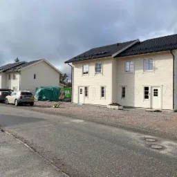 Dwa identyczne, dwukondygnacyjne budynki w trakcie budowy z elewacją z jasnego drewna i czarnym dachem, zaparkowane samochody i kontener na gruz w tle.