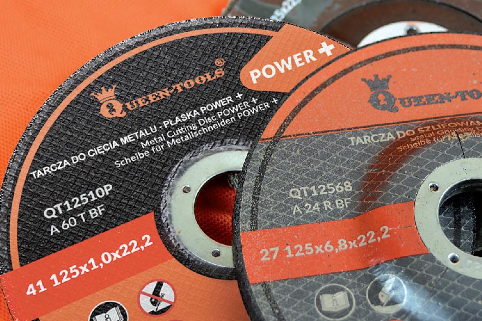 Dwie tarcze do cięcia i szlifowania metalu marki Queen-Tools, z widocznymi oznaczeniami technicznymi i ostrzeżeniami, ułożone na pomarańczowym tle.