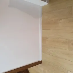 Jasne panele podłogowe ułożone wzdłuż ściany pomalowanej na jasny kolor z białymi listwami przypodłogowymi i ciemnobrązową listwą podłogową.