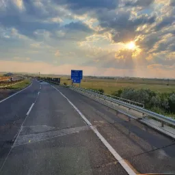 Autostrada w słoneczny dzień. Widok z kabiny, tablica informacyjna z odległościami do miast. Krajobraz z polami i zachmurzonym niebem w tle.