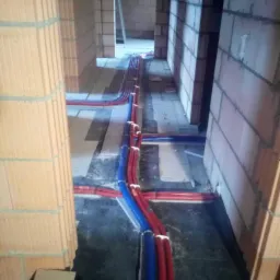 Rozprowadzone rury ogrzewania podłogowego w surowym stanie, czerwone i niebieskie, widoczne ściany z cegieł i kartonowe pudełko.