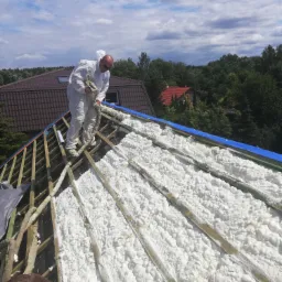 Pracownik w kombinezonie ochronnym aplikuje piankę izolacyjną na poddaszu w Warszawie, widoczne drewniane belki konstrukcyjne i fragment nieba z chmurami.