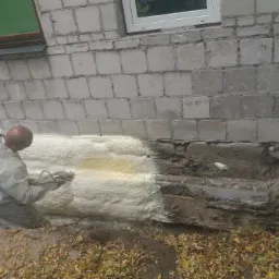 Ocieplanie fundamentów pianką PUR: pracownik w kombinezonie nanosi izolację na mur z pustaków, widoczne fragmenty nieocieplonej ściany i wystające rury.