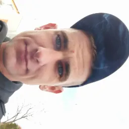 Portret mężczyzny z niebieskimi oczami w czapce bejsbolówce z widoczną częścią drzewa w tle.