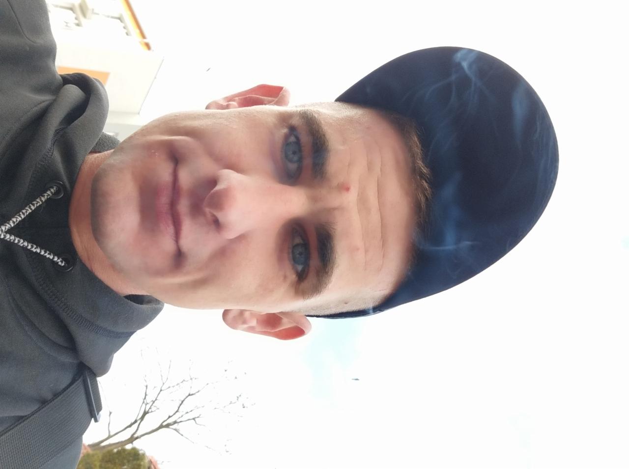 Portret mężczyzny z niebieskimi oczami w czapce bejsbolówce z widoczną częścią drzewa w tle.