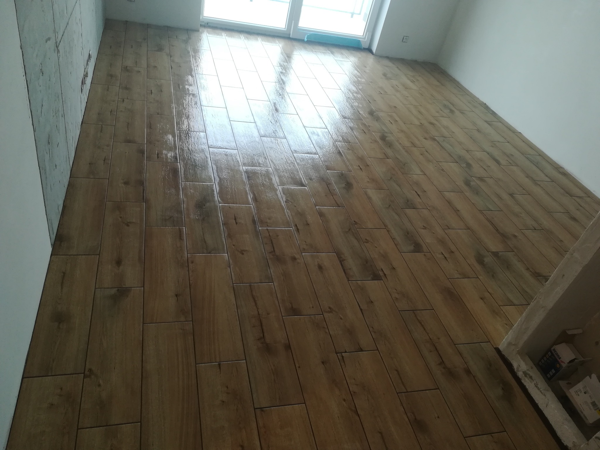 Ułożone, świeżo wyczyszczone płytki podłogowe imitujące drewno w jasnym odcieniu, widoczne fugi i fragment ściany w stanie surowym.