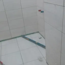 Świeżo wykafelkowana łazienka z poziomnicą na podłodze, widoczne wystające rury z instalacji wodnej, szare płytki na ścianach i podłodze, etap prac wykończeniowych.