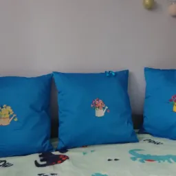 Trzy niebieskie poduszki dekoracyjne z haftowanymi wzorami konewek z kwiatami, ułożone na dziecięcej pościeli z motywem dinozaurów.