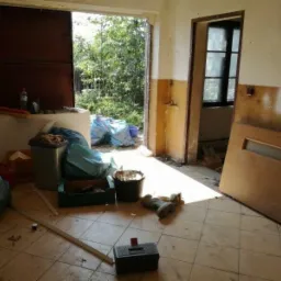 Wnętrze zniszczonego pomieszczenia w trakcie opróżniania, z workami na śmieci, wiadrem, skrzynką narzędziową i otwartymi drzwiami na zewnątrz.