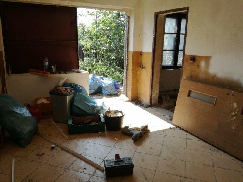 Wnętrze zniszczonego pomieszczenia w trakcie opróżniania, z workami na śmieci, wiadrem, skrzynką narzędziową i otwartymi drzwiami na zewnątrz.