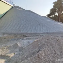 Skarpa zabezpieczona betonową kostką o strukturze plastra miodu, widoczny stos piasku i paleta z kostką, w tle drzewa i fragment budynku.