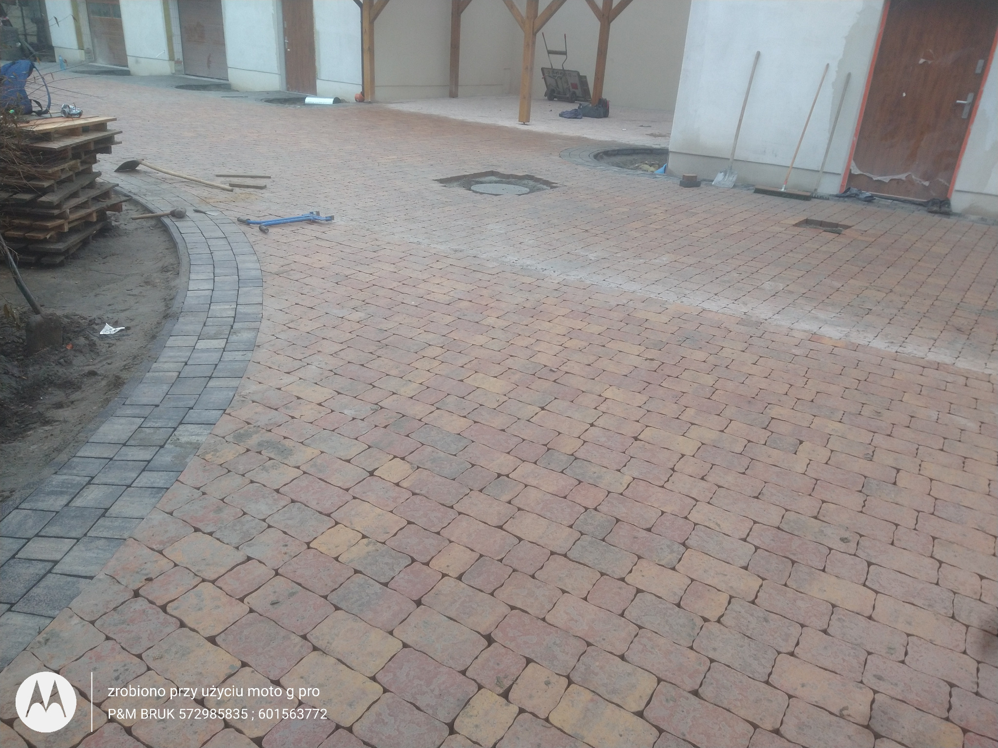Częściowo ułożona nawierzchnia z kostki brukowej w odcieniach brązu i szarości, widoczne narzędzia brukarskie, paleta z kostką, oraz przygotowane podłoże pod obrzeża.