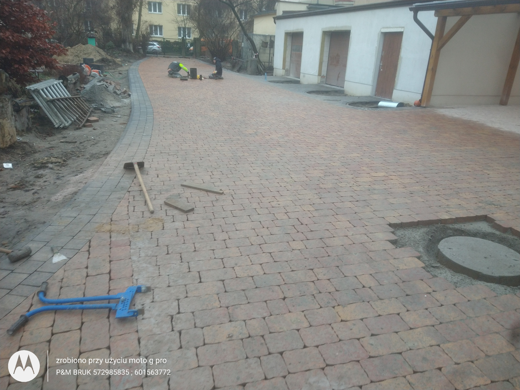 Trwa układanie beżowo-brązowej kostki brukowej na posesji, widoczne narzędzia brukarskie, fragment budynku w tle i dwóch pracowników przy pracy.