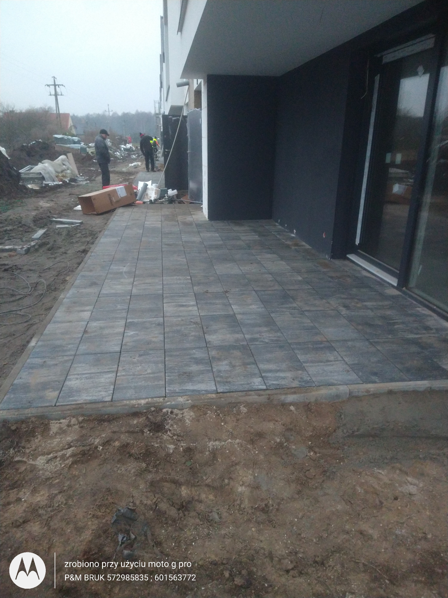 Ułożona szara kostka brukowa przed nowoczesnym budynkiem z czarną elewacją, widoczni pracownicy w oddali, teren wokół w trakcie prac ziemnych.