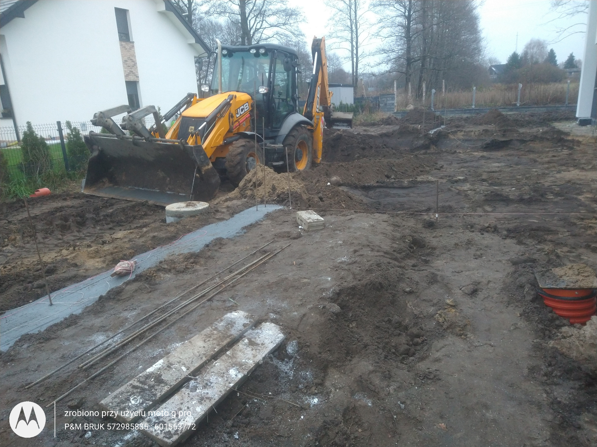 Żółta koparko-ładowarka JCB na placu budowy, przygotowująca teren pod przyszłą nawierzchnię. Widoczne wytyczone obszary, rury kanalizacyjne i materiały budowlane.