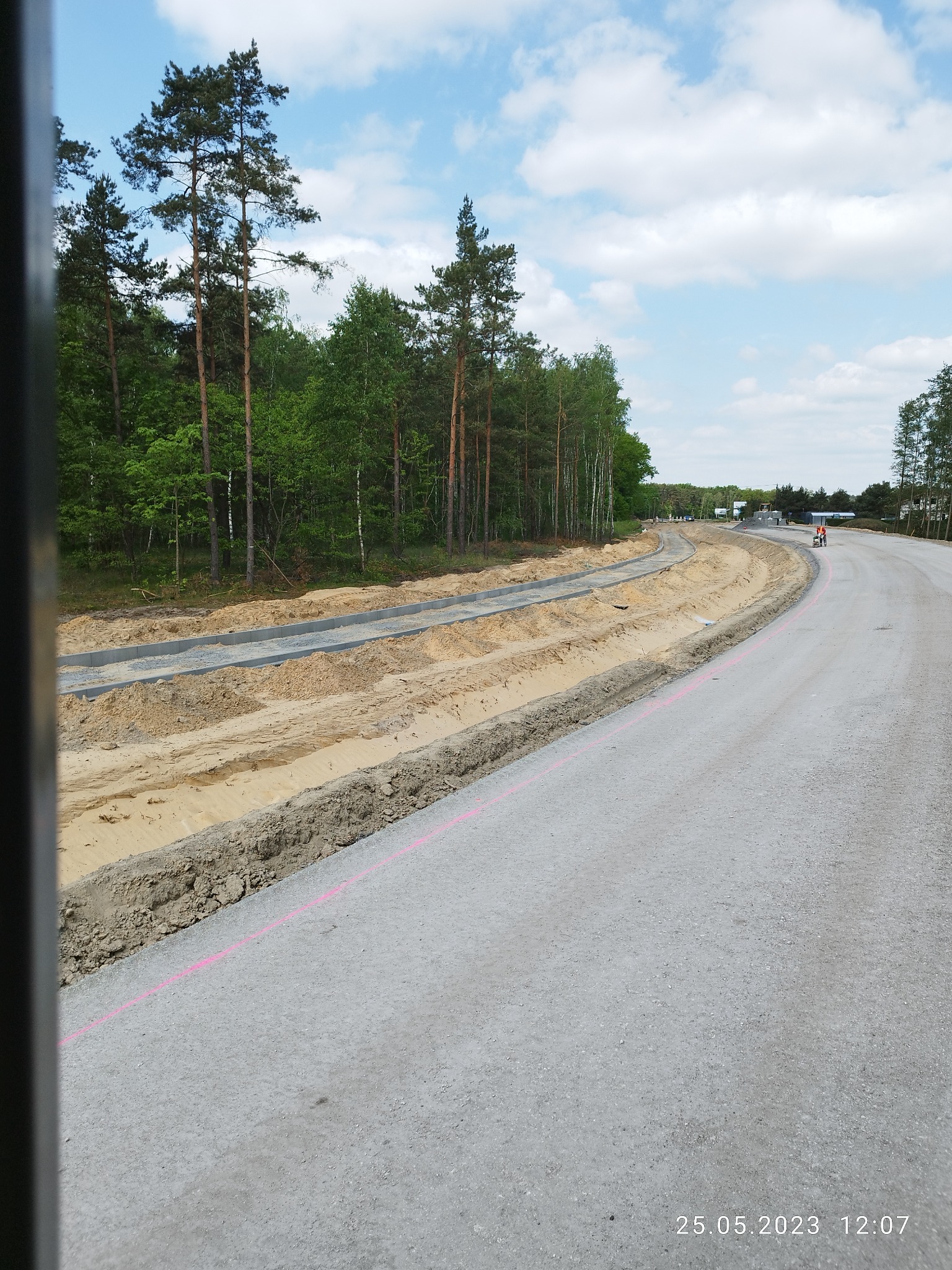 Wyrównywanie terenu i układanie krawężników wzdłuż nowej drogi, widoczne różowe oznaczenia geodezyjne, w tle las.