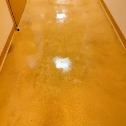 Błyszcząca, pomarańczowa posadzka w korytarzu, świeżo umyta, z widocznymi refleksami światła, sfotografowana z perspektywy podłogi, z fragmentem drzwi po lewej stronie.