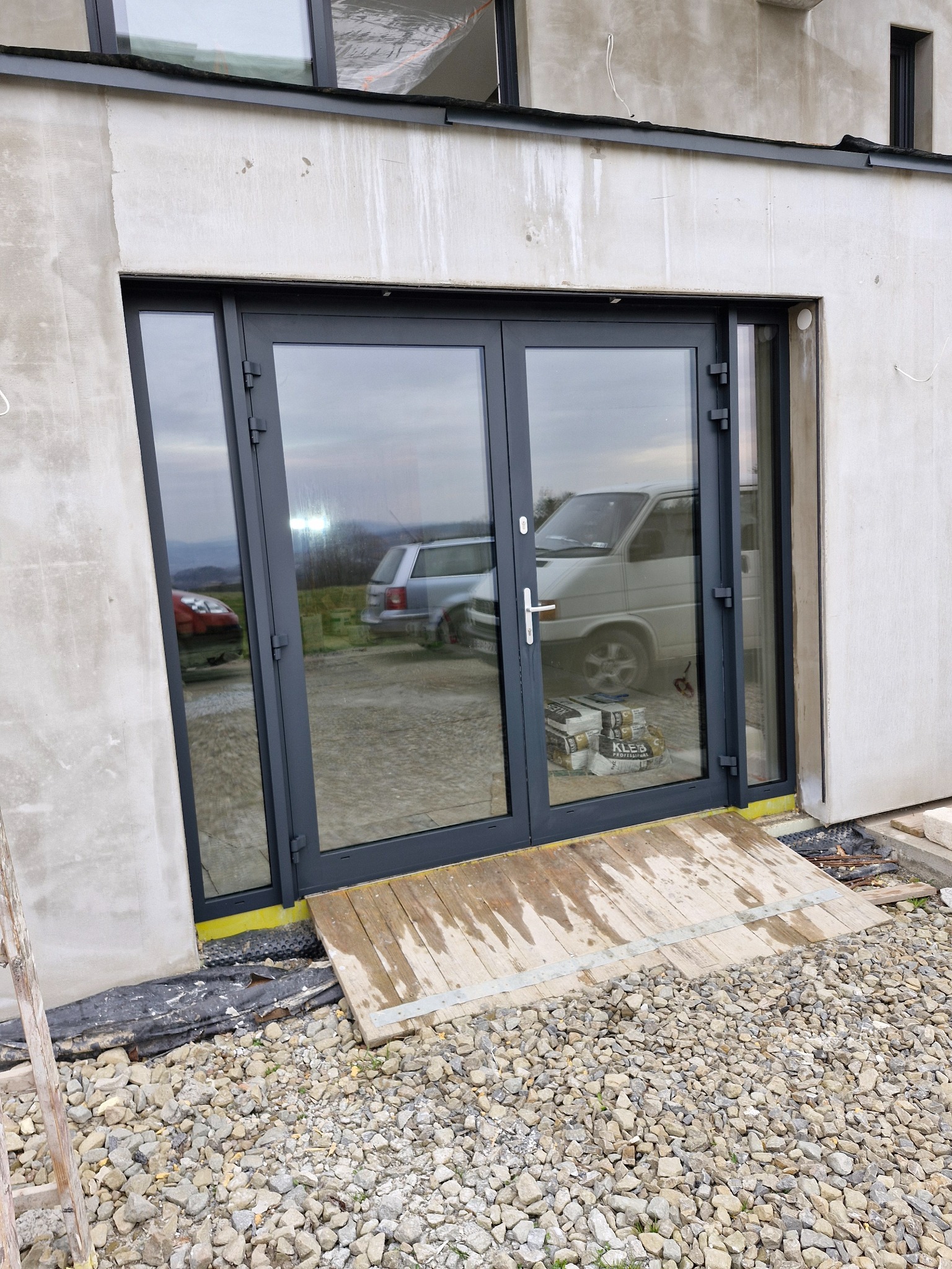 Nowe, antracytowe drzwi tarasowe z widocznymi refleksami otoczenia, zamontowane w surowym budynku z widoczną izolacją i tymczasowym podjazdem z desek.