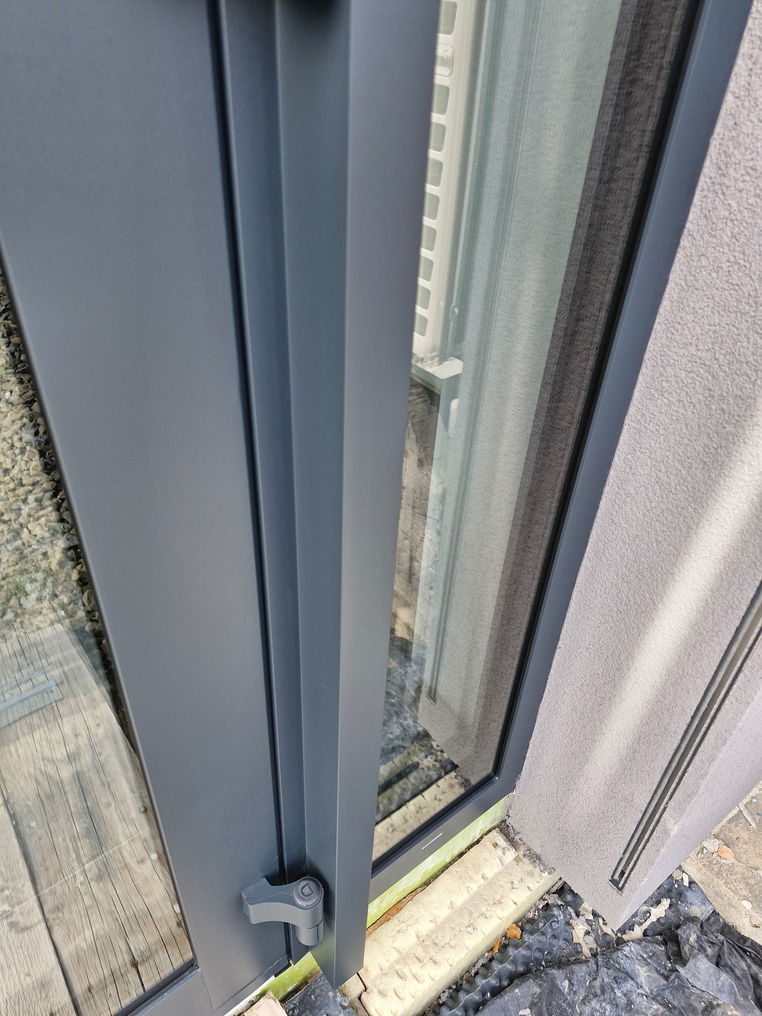 Szczegółowe ujęcie nowej, szarej ramy okiennej z widoczną klamką i fragmentem elewacji budynku z fakturą oraz widokiem na zewnątrz przez szybę.