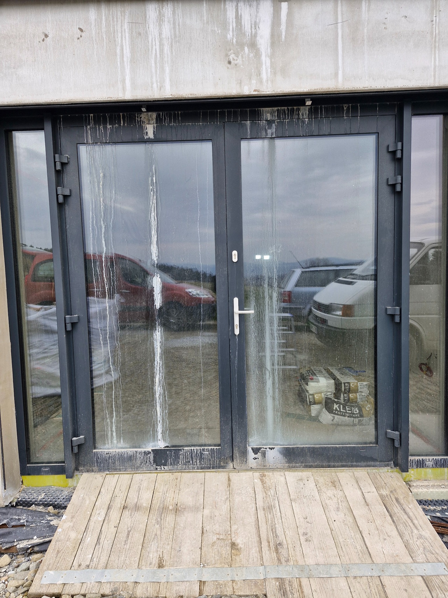 Brudne, przeszklone drzwi balkonowe z zaciekami po remoncie, widoczne worki z klejem wewnątrz, drewniany podest wejściowy, w tle zaparkowane samochody.