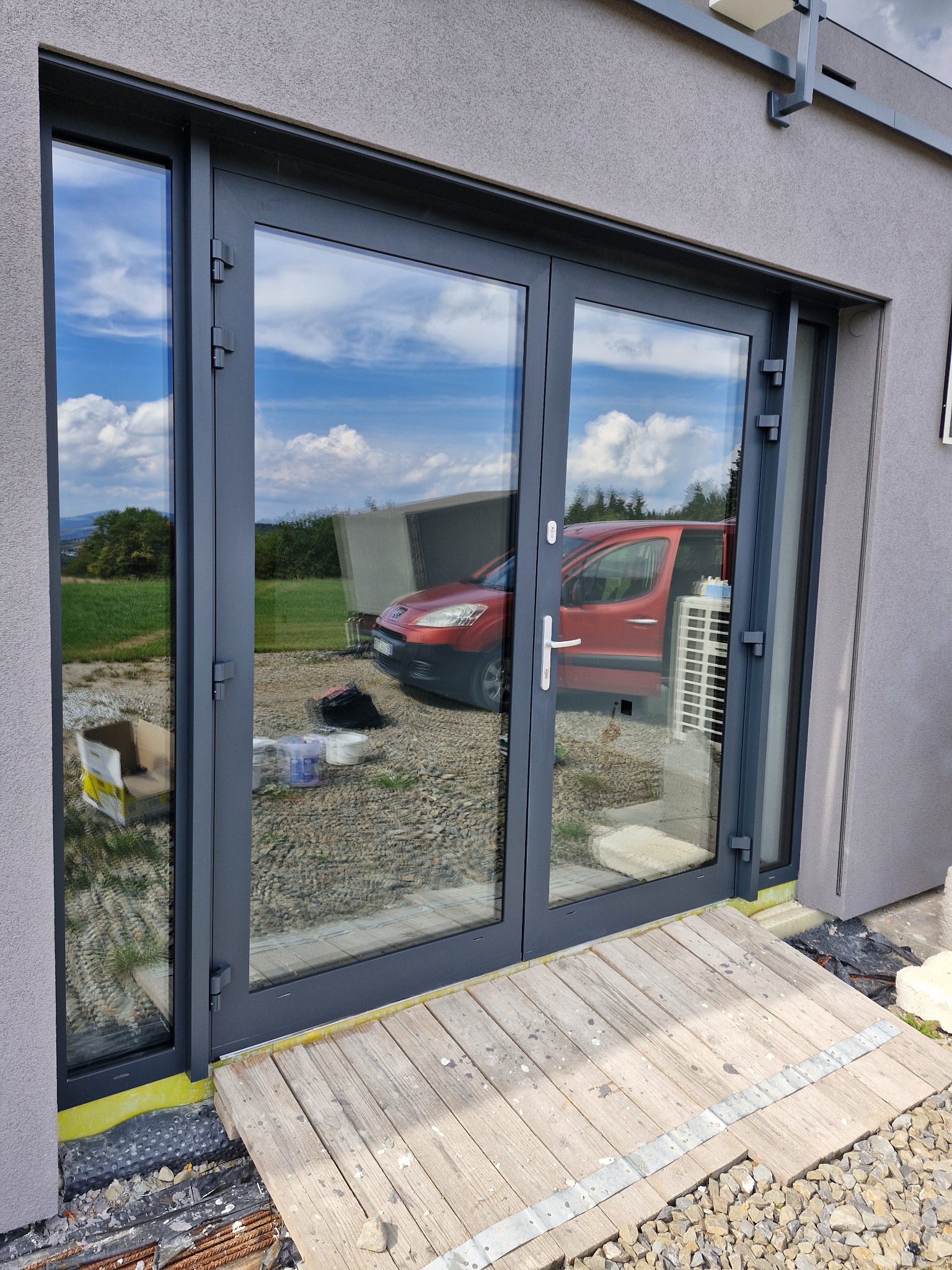 Szare, dwuskrzydłowe drzwi balkonowe z widocznym odbiciem nieba, trawnika i czerwonego samochodu, zamontowane w szarej elewacji budynku, z drewnianym podestem u dołu i widocznymi elementami...