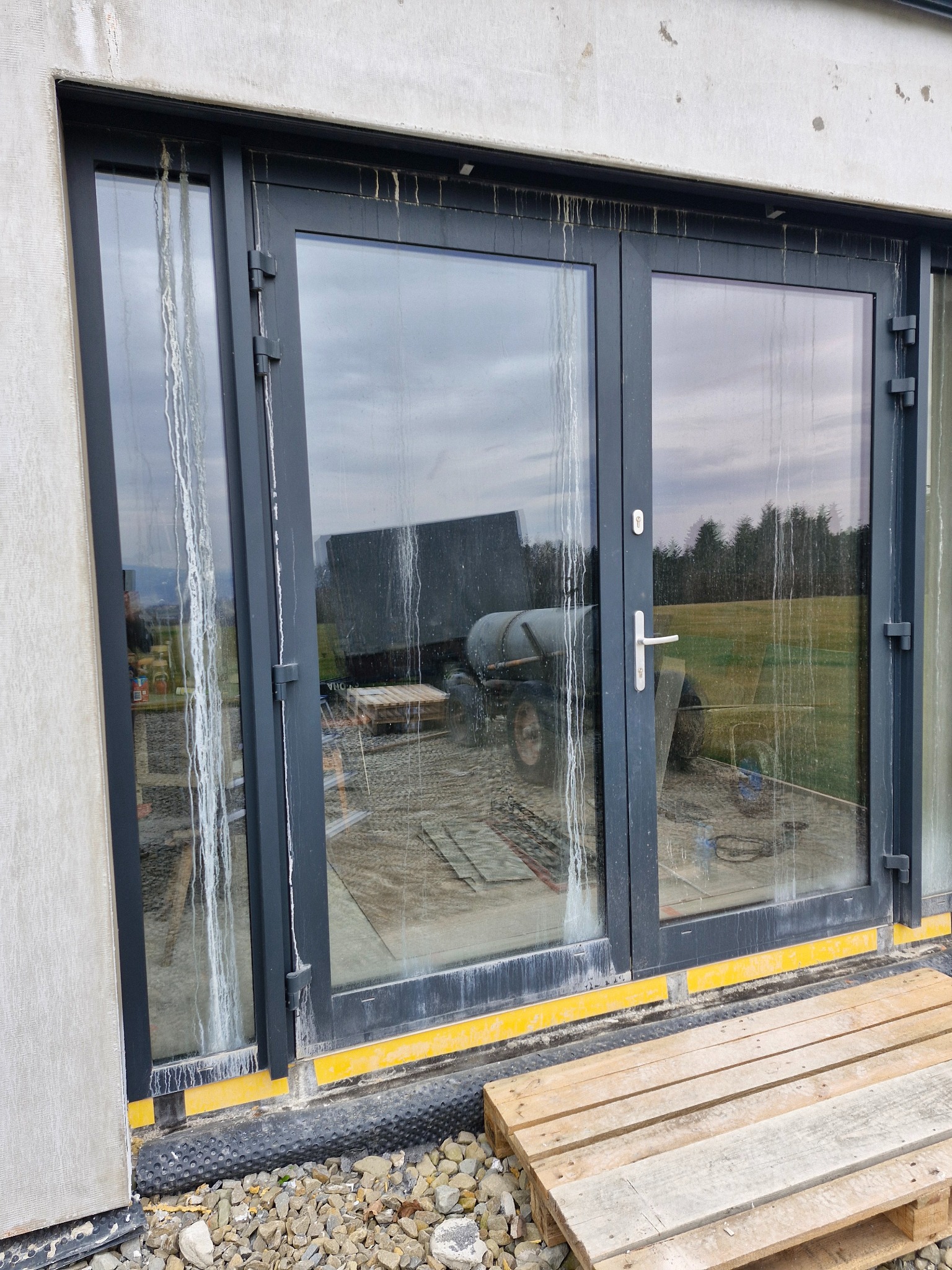 Brudne okna tarasowe z ciemnymi ramami po remoncie, widoczne zacieki i kurz, na zewnątrz traktor z przyczepą, paleta drewniana.