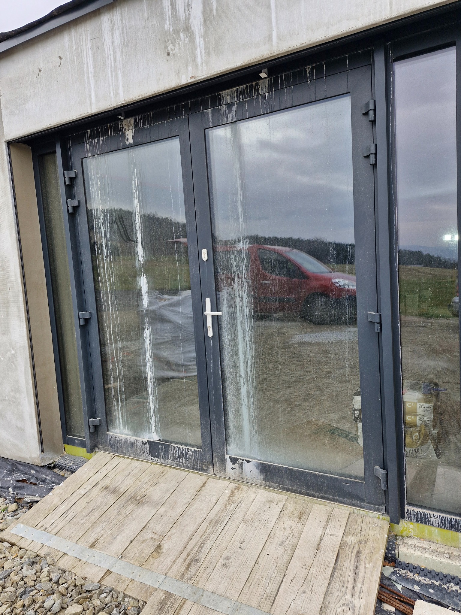 Brudne, przeszklone drzwi tarasowe z czarnymi ramami, z zaciekami po budowie, prowadzące na drewniany podest, w tle czerwony samochód i teren budowy.