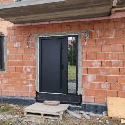 TYLKA Okna Drzwi Bramy Rolety Pergole i Ogrodzenia - Ciemna, nowoczesna brama wejściowa zamontowana w surowym stanie budynku z czerwonej cegły, widoczne przewody i tymczasowe schody z palet.