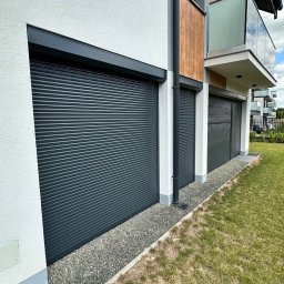 TYLKA Okna Drzwi Bramy Rolety Pergole i Ogrodzenia - Nowoczesny dom z trzema szarymi bramami garażowymi rolowanymi, obok balkonu z szklaną balustradą. Widok z zewnątrz, trawnik i kamień ozdobny u podstawy.