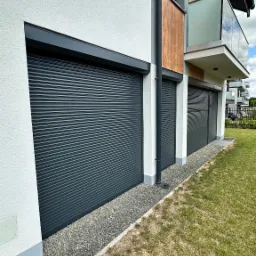 Nowoczesny dom z trzema szarymi bramami garażowymi rolowanymi, obok balkonu z szklaną balustradą. Widok z zewnątrz, trawnik i kamień ozdobny u podstawy.