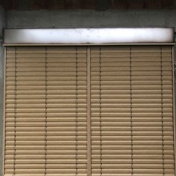 TYLKA Okna Drzwi Bramy Rolety Pergole i Ogrodzenia - Zamknięte, beżowe żaluzje fasadowe na tle surowej ściany z betonu i cegieł. Widoczne elementy montażowe i fragment trawy u dołu zdjęcia.