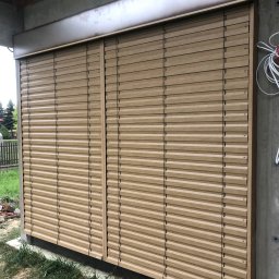 TYLKA Okna Drzwi Bramy Rolety Pergole i Ogrodzenia - Zamknięte żaluzje fasadowe w kolorze drewna, zamontowane na zewnątrz budynku w trakcie budowy. Widoczne przewody elektryczne i elementy konstrukcyjne.