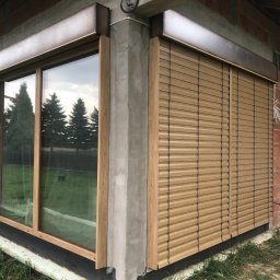 TYLKA Okna Drzwi Bramy Rolety Pergole i Ogrodzenia - Drewniane żaluzje fasadowe zamontowane na oknie budynku w trakcie budowy. Widoczne niedokończone ściany z cegły i styropian. Drewniana rama okna i szare elementy konstrukcyjne.