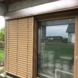 Zamknięta, beżowa roleta zewnętrzna zamontowana obok okna z drewnianą ramą w surowym stanie budynku. W tle widok na zielone pole i dom.