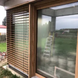 Zamknięte żaluzje drewniane w oknie z drewnianą ramą na tle trawnika i budynków. Widoczna drabina oparta o szybę okna.