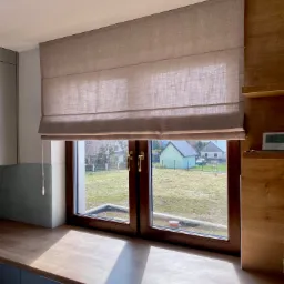 Beżowa roleta rzymska zamontowana w oknie z brązowymi ramami, widok na zielony ogród i domy, blat kuchenny z połyskującym światłem słonecznym.