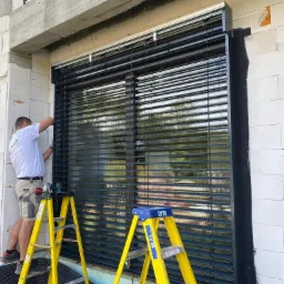 Montaż czarnych żaluzji wewnętrznych przez instalatora na żółtej drabinie w surowym stanie budynku.