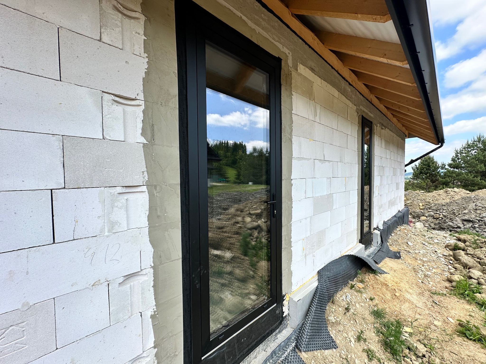 Czarna rama okienna w surowym murze z betonu komórkowego, widoczne ocieplenie fundamentów i drewniana konstrukcja dachu. Okno z widokiem na zieleń.