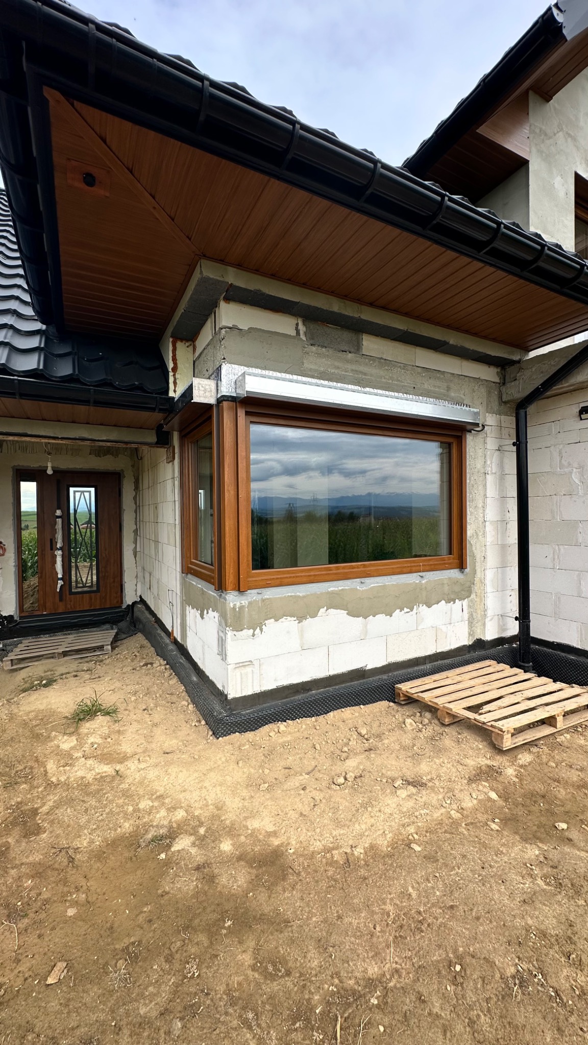 Brązowe okno PCV z roletą zewnętrzną w domu w trakcie budowy. Widok na góry przez szybę. Paleta drewniana na ziemi. Czarna rynna i podbitka dachowa.