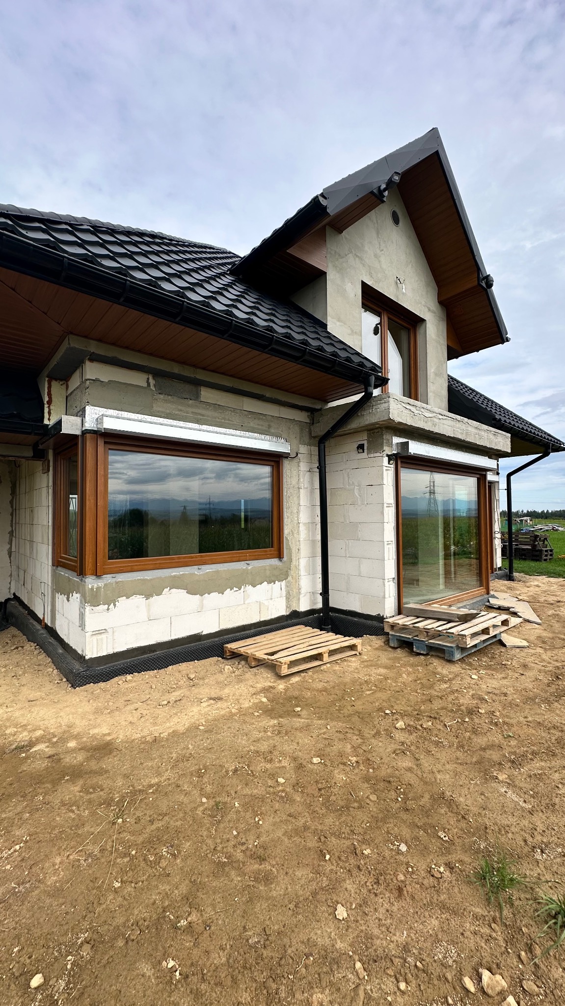 Surowy stan domu jednorodzinnego z drewnianymi oknami i przygotowaną instalacją rolet zewnętrznych. Budynek w trakcie wykończenia, widoczne palety budowlane.