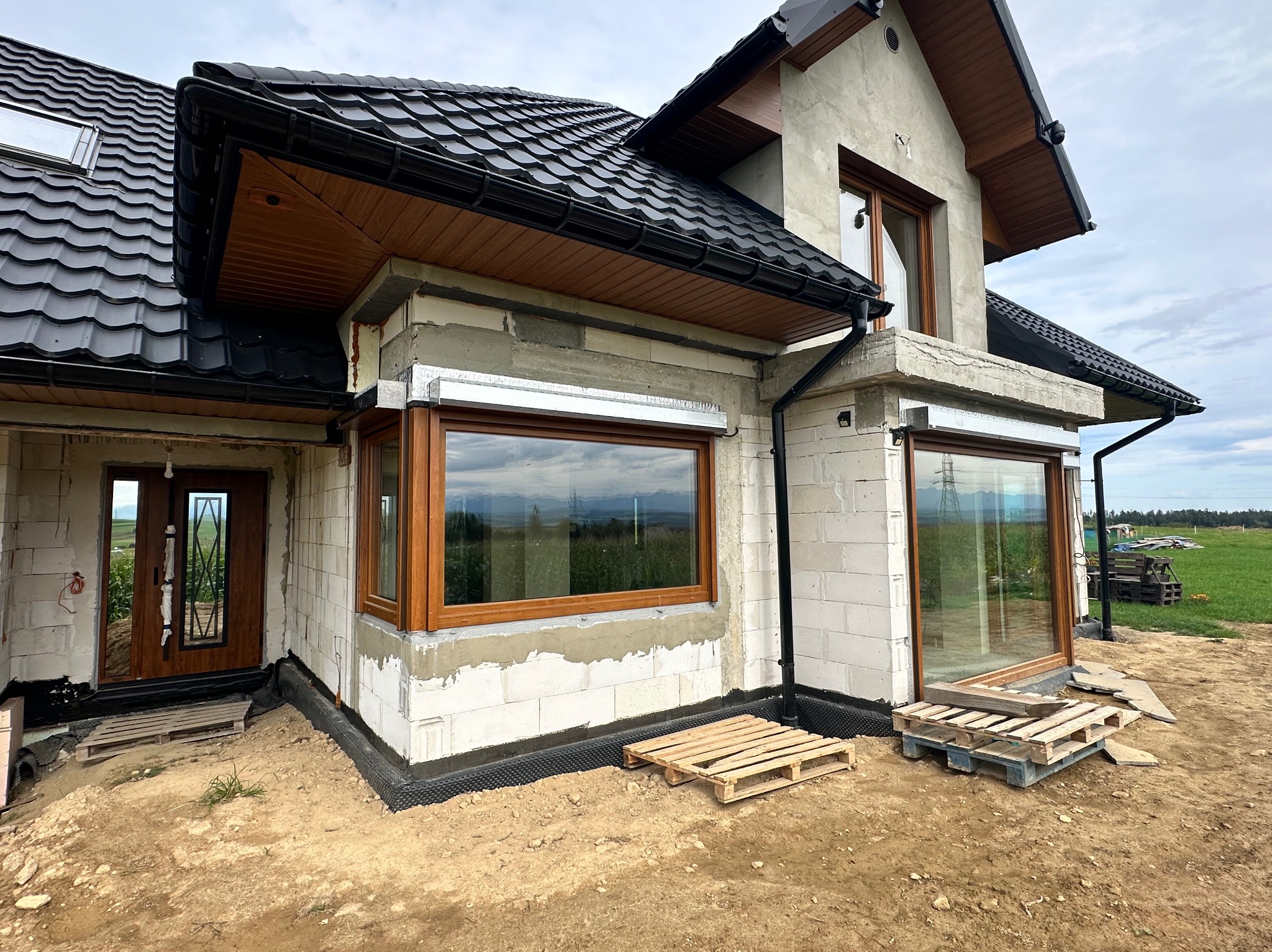 Dom w trakcie budowy z drewnianymi oknami i drzwiami. Widoczne bloczki betonowe, czarna dachówka i palety. Na zewnątrz pole i pochmurne niebo.