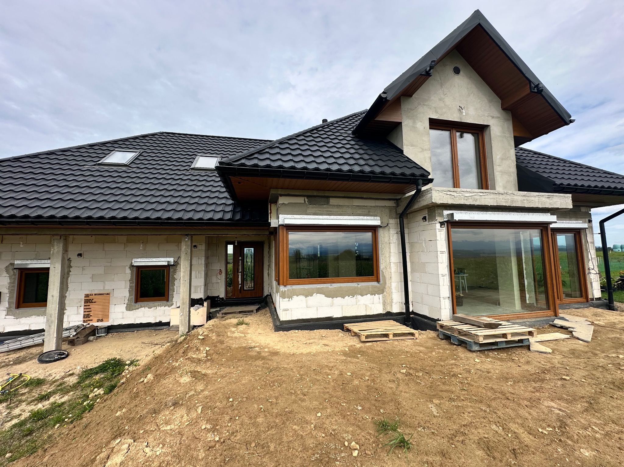 Dom w stanie surowym otwartym z brązowymi oknami i czarnym dachem. Widoczne palety i ziemia wokół budynku. Okna częściowo z roletami zewnętrznymi.