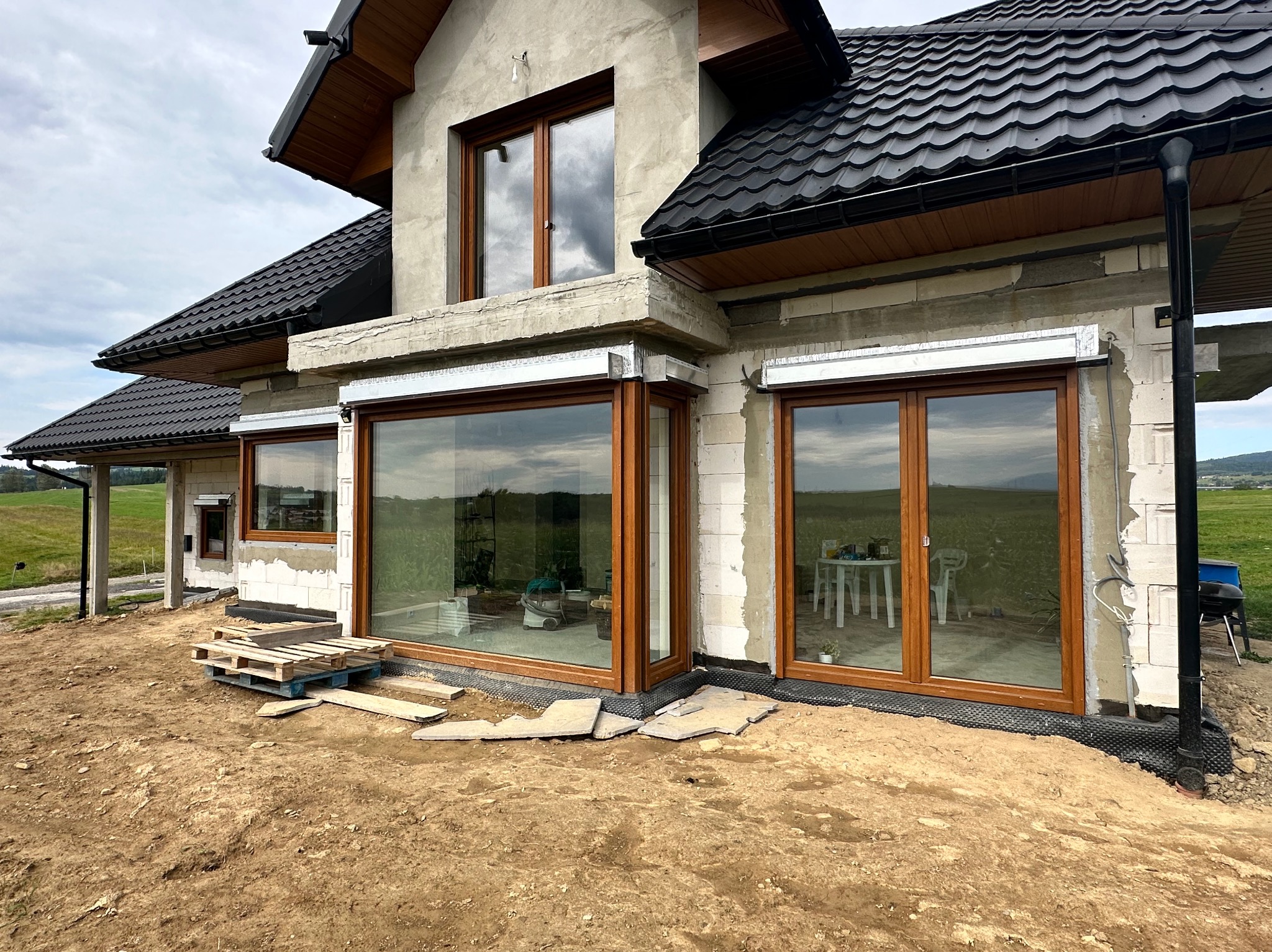 Surowy stan domu jednorodzinnego z brązowymi oknami i przygotowanym podłożem. Widoczne palety i ziemia. Okna z roletami zewnętrznymi w srebrnej obudowie.