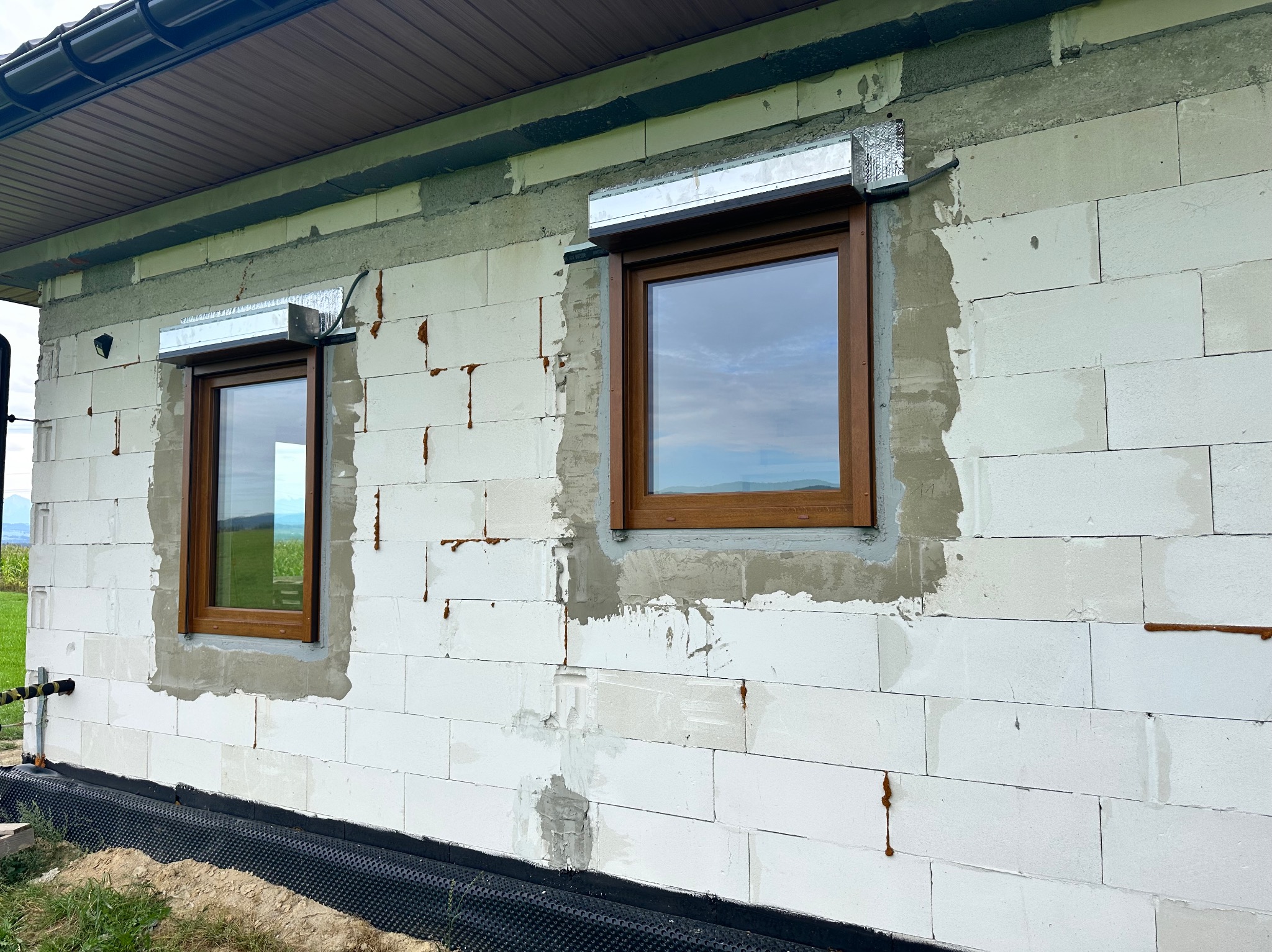 Dwa brązowe okna PCV z roletami zewnętrznymi, zamontowane w ścianie z białych bloczków. Widoczne ślady po montażu i fragment dachu.