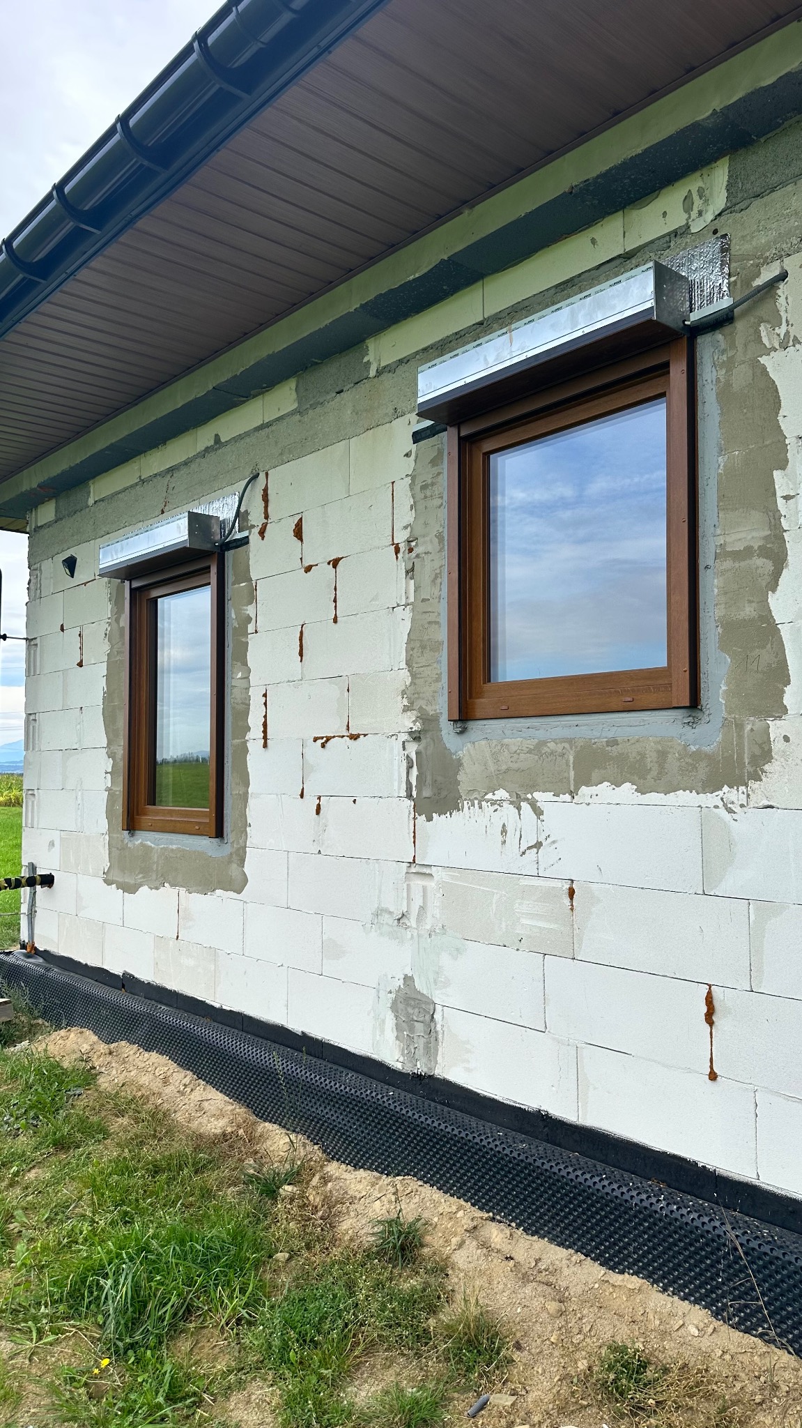 Montaż okien PCV z brązową ramą i roletami zewnętrznymi na surowej ścianie budynku z betonu komórkowego. Widoczne detale montażu.